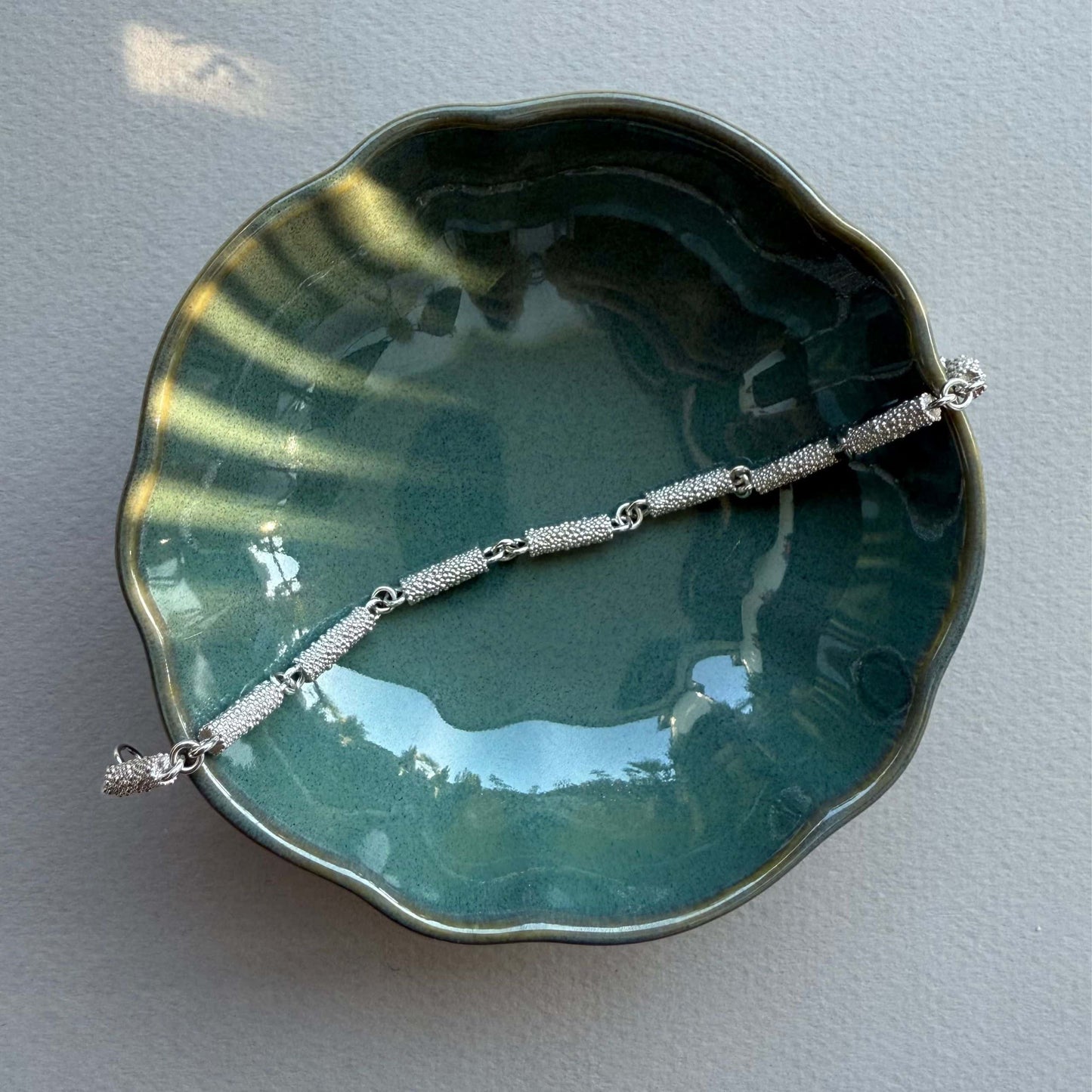 Armband aus Fairmined Silber mit fein texturierter Oberfläche, arrangiert in kleinem Keramikschälchen bei natürlichem Licht