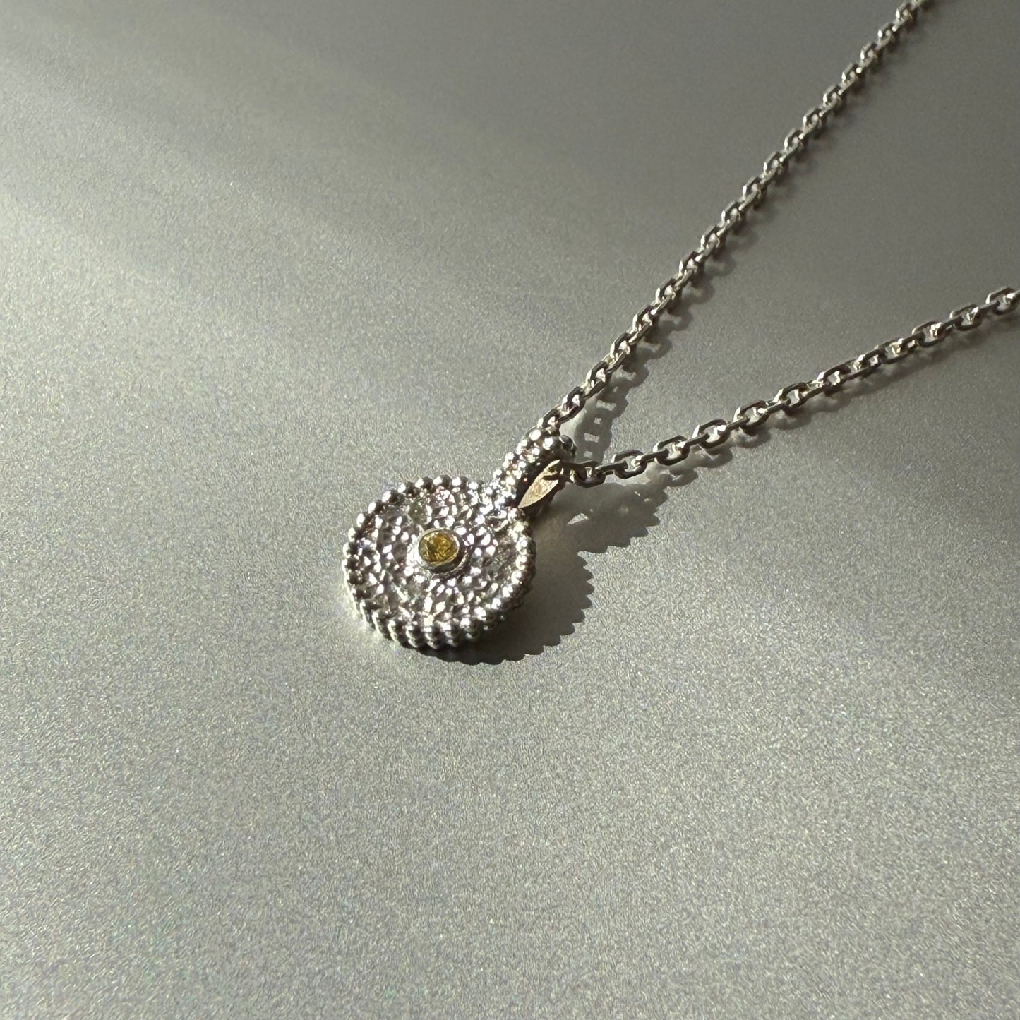 Nahaufnahme eines runden Anhängers aus Fairmined Silber mit hellgelbem, fair gehandeltem Saphir, liegend auf silbernem Untergrund bei natürlichem Licht