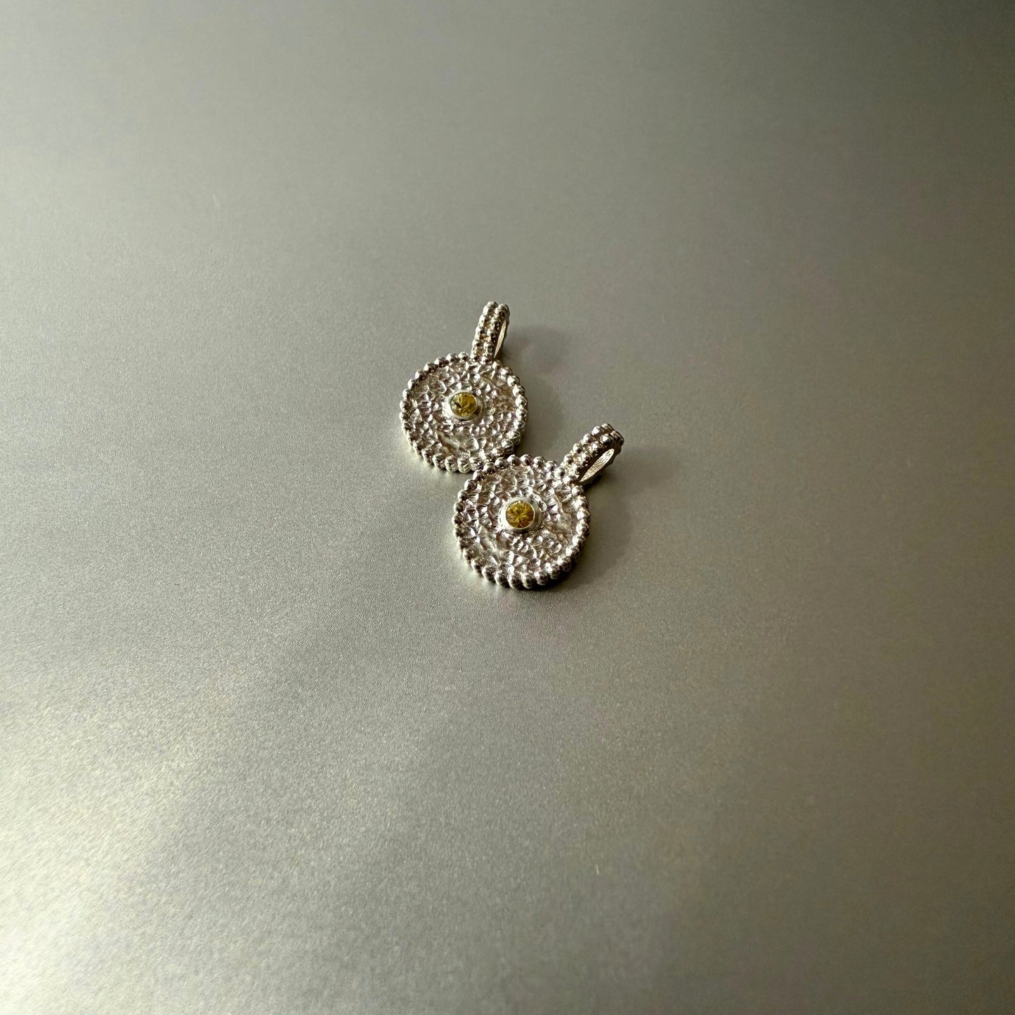 Zwei runde Anhänger aus Fairmined Silber mit hellgelbem, fair gehandeltem Saphir, nebeneinander liegend auf silbernem Untergrund bei natürlichem Licht