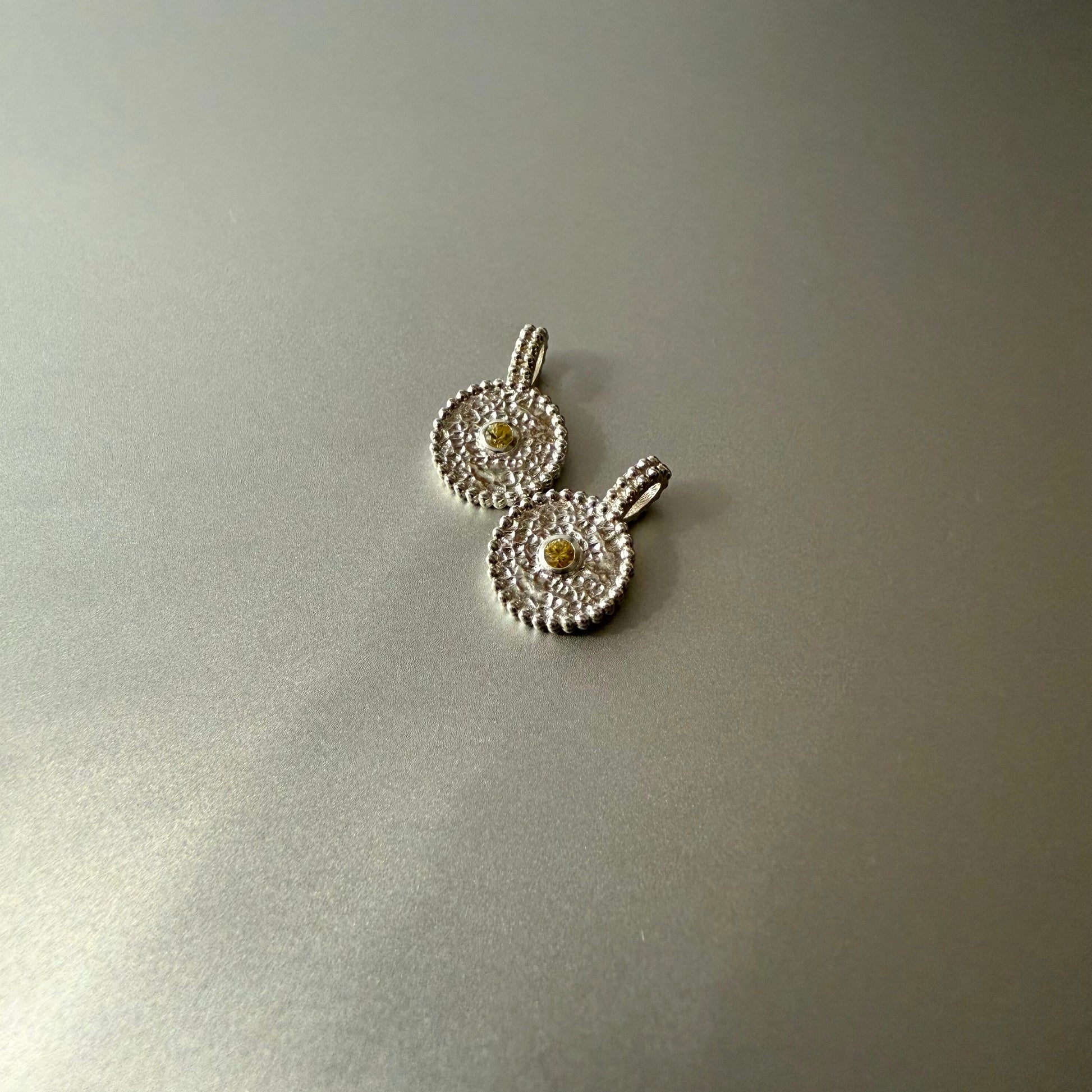 Zwei runde Anhänger aus Fairmined Silber mit hellgelbem, fair gehandeltem Saphir, nebeneinander liegend auf silbernem Untergrund bei natürlichem Licht