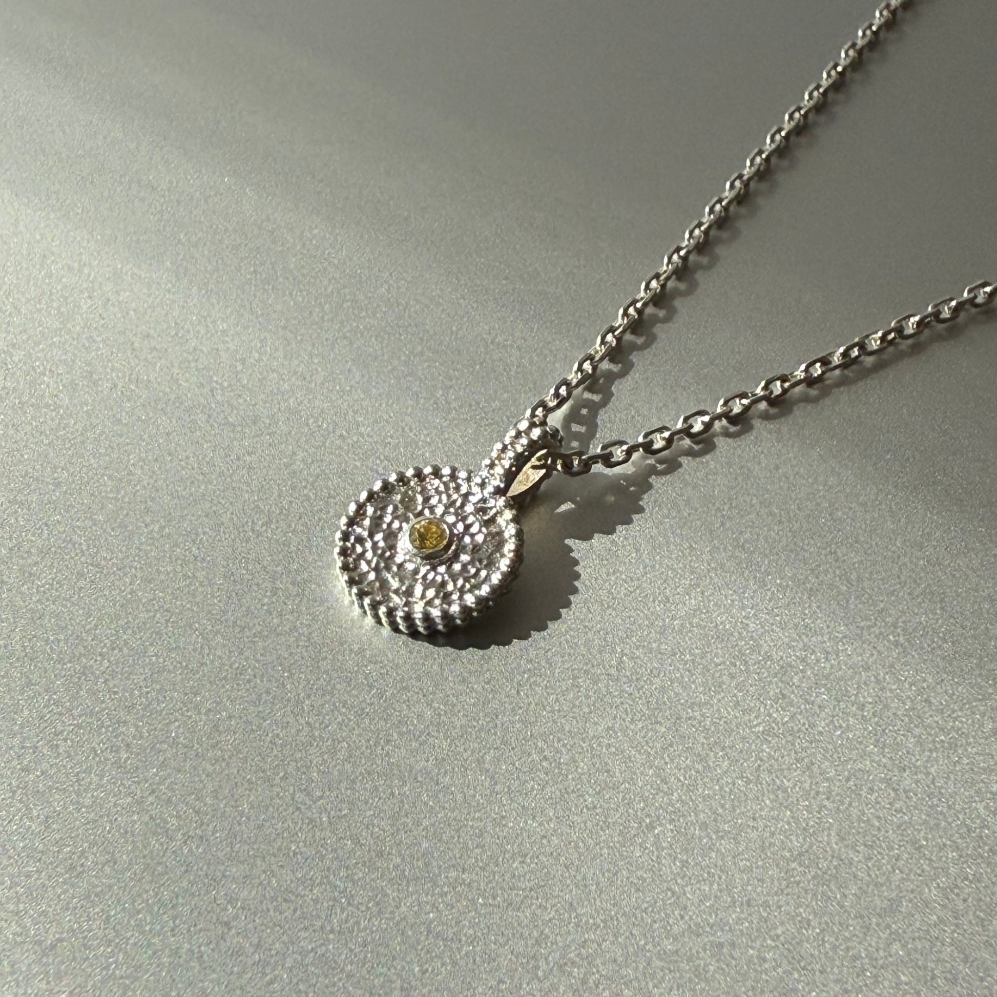 Nahaufnahme eines runden Anhängers aus Fairmined Silber mit hellgelbem, fair gehandeltem Saphir, liegend auf silbernem Untergrund bei natürlichem Licht