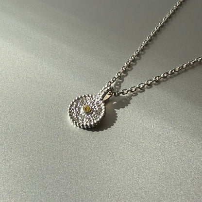 Nahaufnahme eines runden Anhängers aus Fairmined Silber mit hellgelbem, fair gehandeltem Saphir, liegend auf silbernem Untergrund bei natürlichem Licht